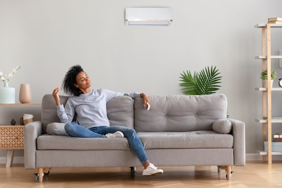 African american relaxed woman sitting on comfortable couch in living room at modern home holds air conditioner remote control enjoying breathing fresh cool air at summer or warm air at winter season (African american relaxed woman sitting.